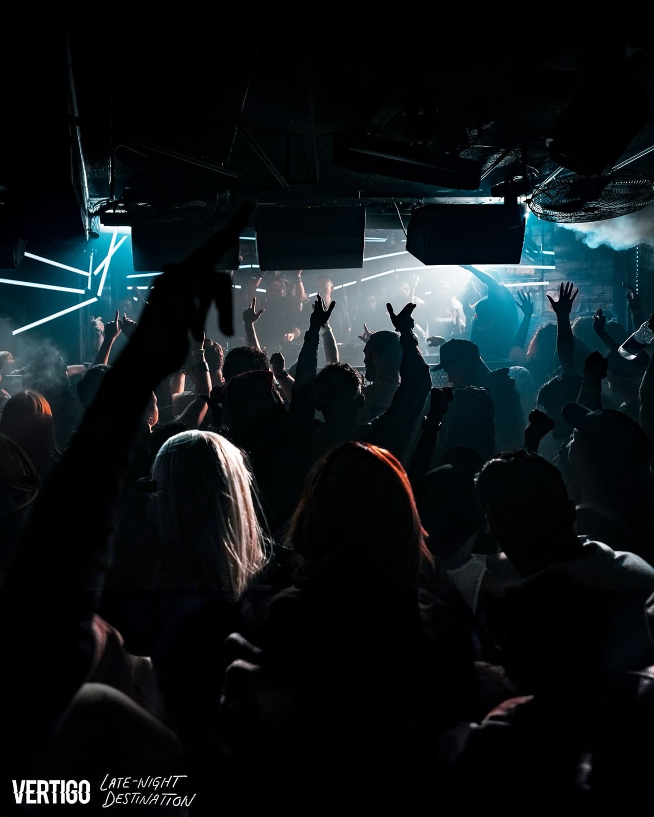 A crowd with raised hands on the dance floor at Vertigo under blue strip lights and haze